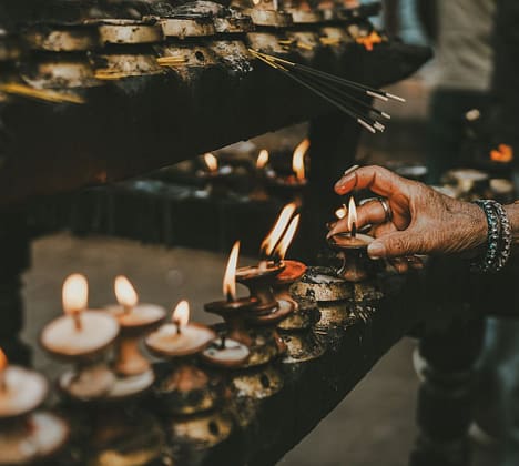 hero-img-02 Hand lighting oil lamps during a traditional ritual with incense sticks, creating a spiritual and serene atmosphere.