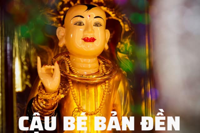 Gilded statue of a boy deity wearing beaded necklaces and bangles with text CẬU BÉ BẢN ĐỀN displayed below.